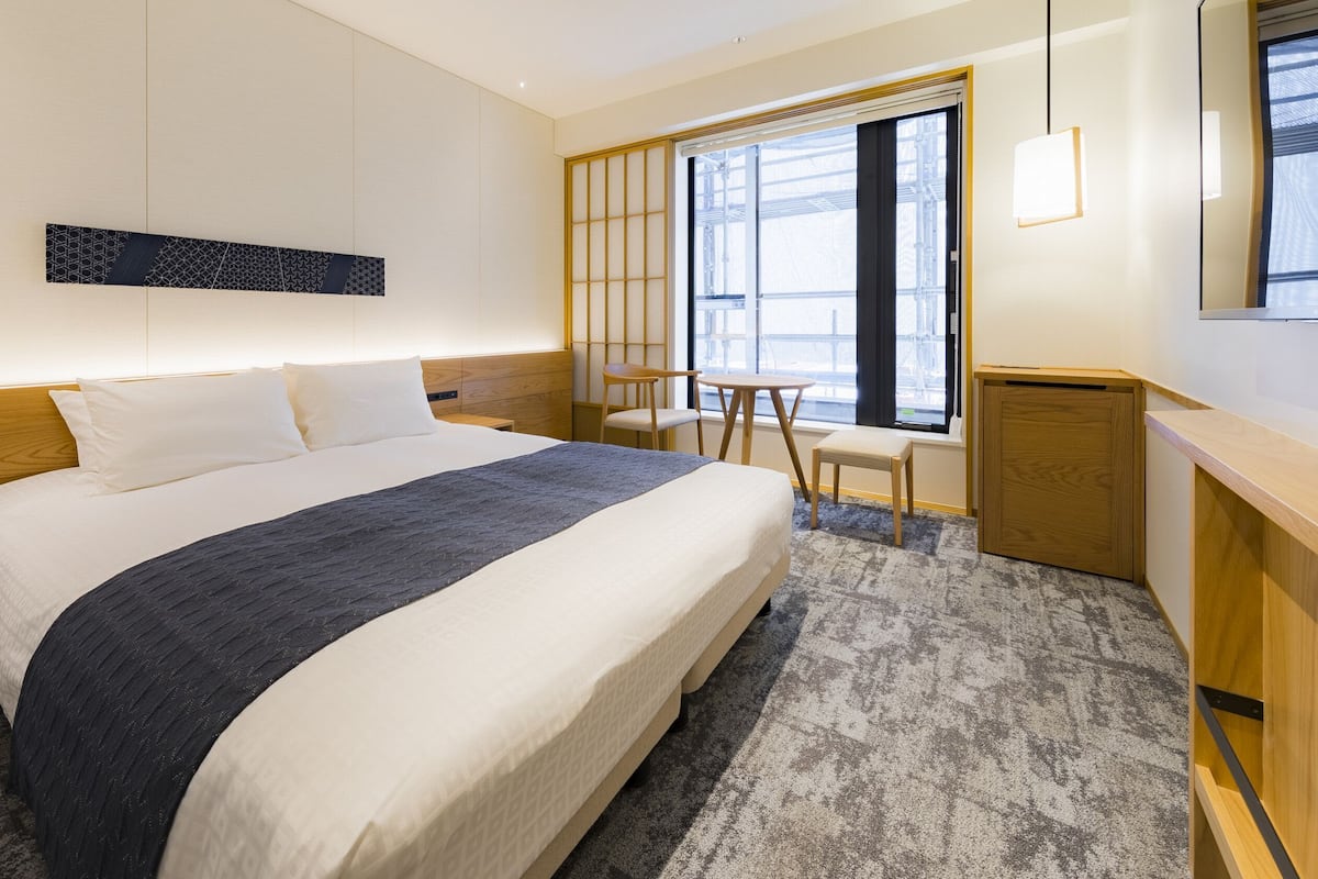 A hotel room with a large bed, bedside tables, a desk, a chair, a window with a view, and a wall-mounted artwork.