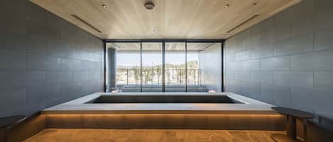 A modern spa area with a large bathtub, a stone floor, and a view of a mountainous landscape through large windows.