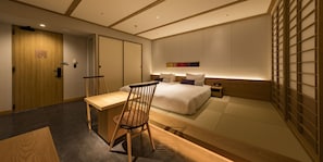 A modern hotel room with a wooden desk, a comfortable chair, a bed with white bedding, and a nightstand.