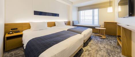 A hotel room with two beds, a small seating area by the window, a desk, and a TV.