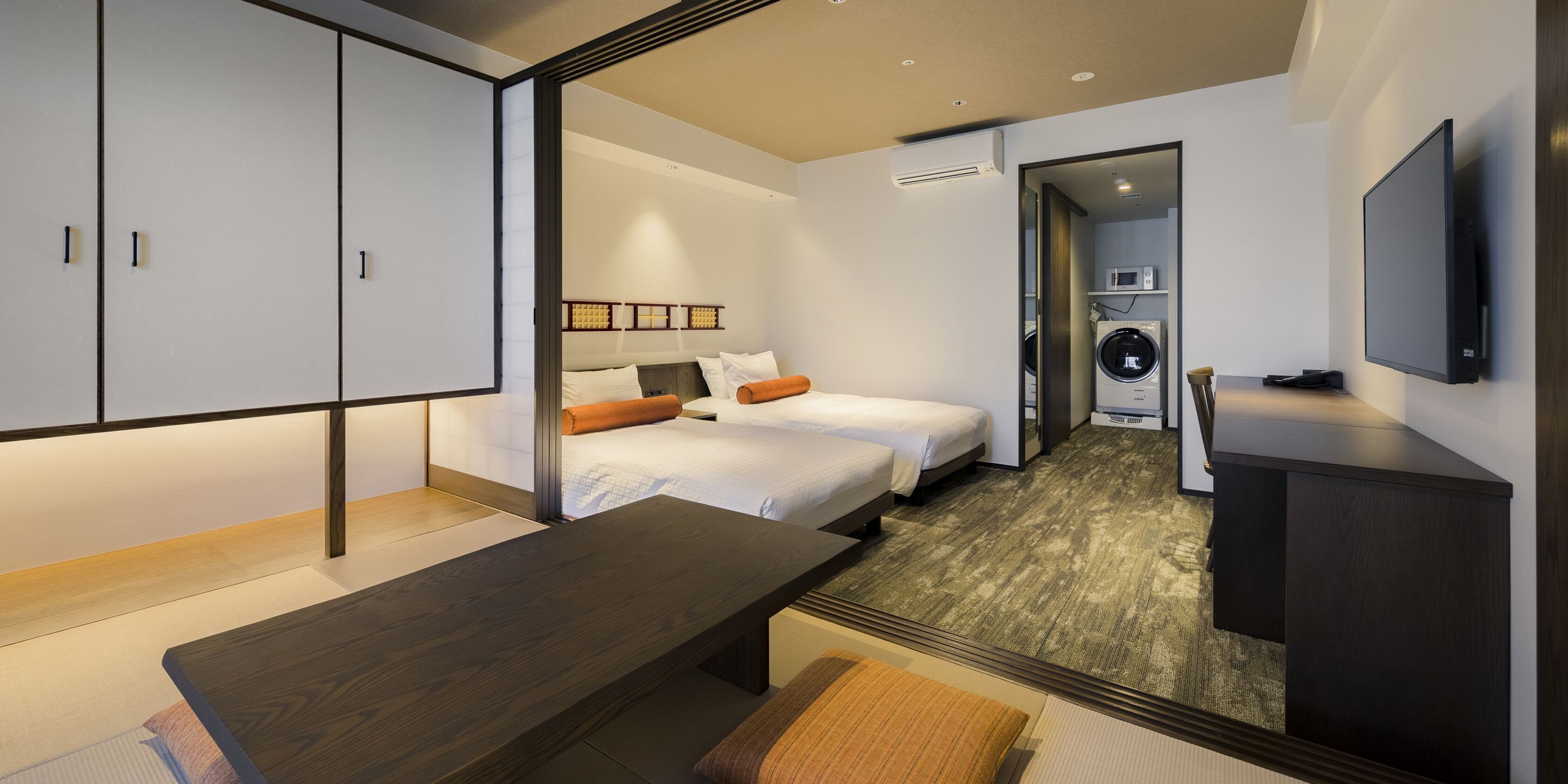 A modern hotel room with a wooden desk, two beds, a washing machine, and a TV.