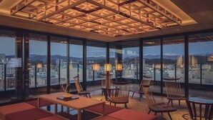 Une pièce spacieuse avec de grandes fenêtres donnant sur la ville, un plafond en bois orné d’un motif géométrique et une table entourée de chaises.
