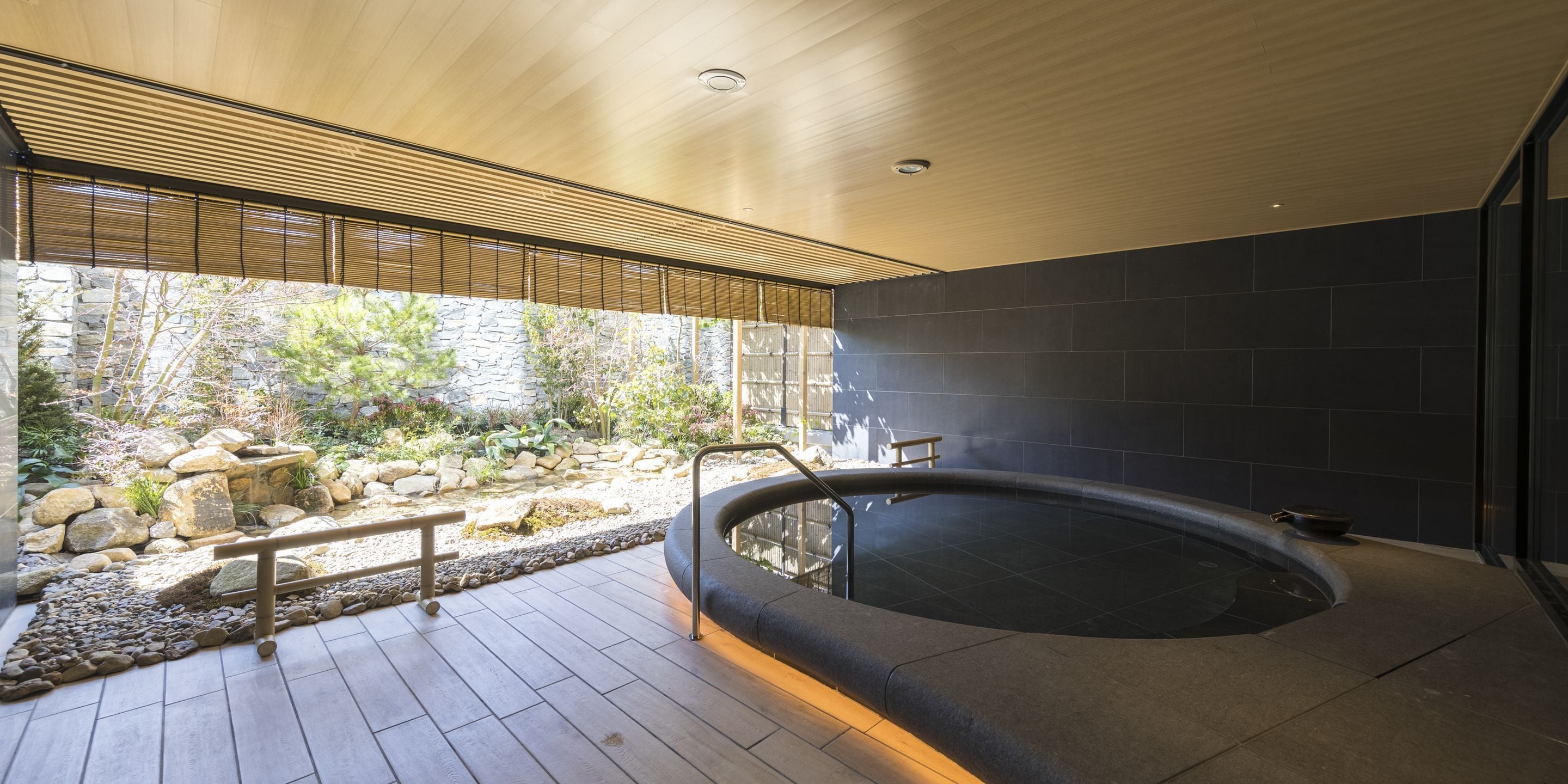 Een moderne spa-ruimte met een jacuzzi, houten terras, stenen muren en uitzicht op een tuin met rotsen en planten.