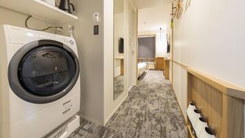 A laundry area with a front-loading washing machine, a small shelf above it, and a hallway leading to a bedroom with a bed and a desk.