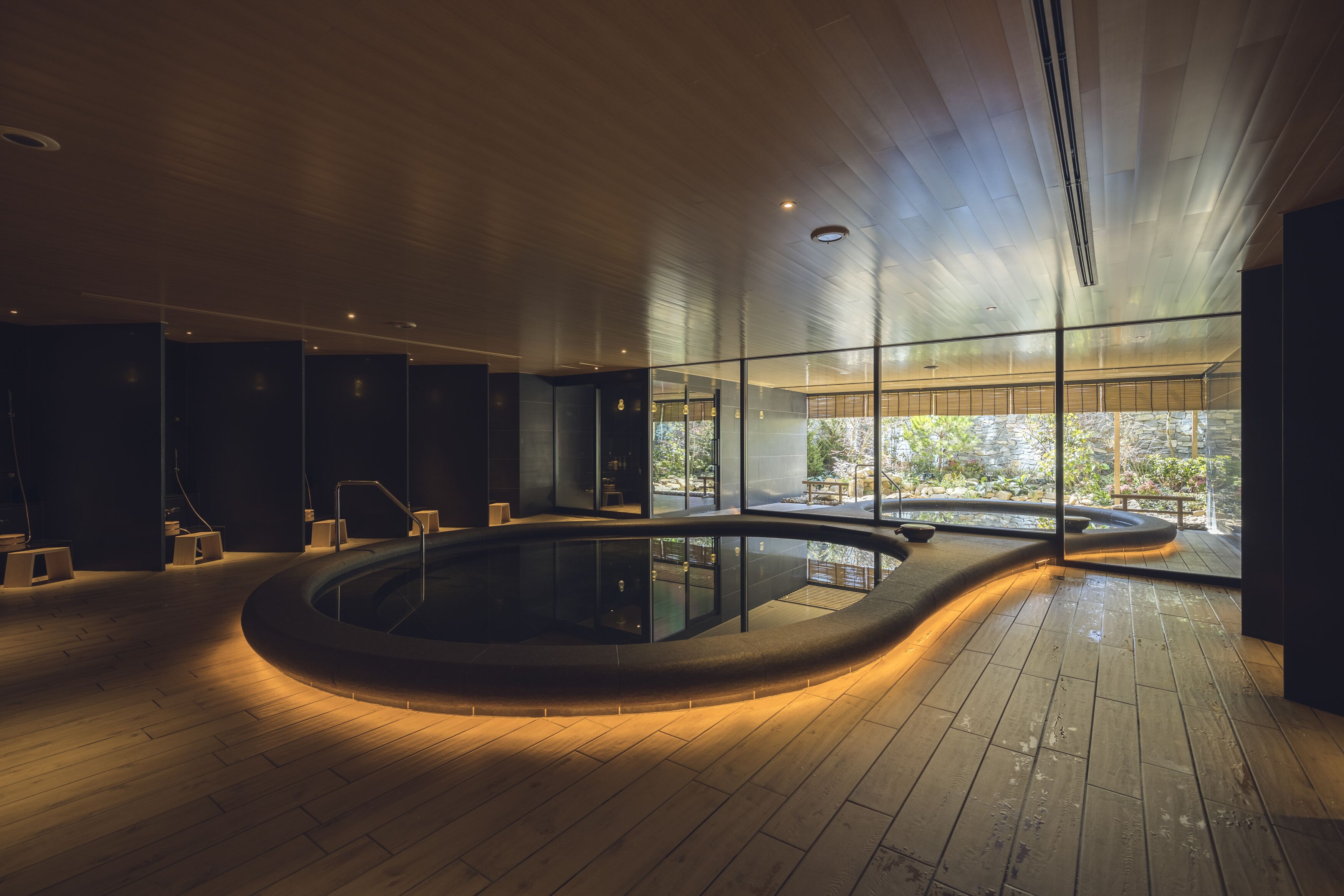 A modern indoor pool area with a wooden floor, glass walls, and a view of greenery outside.