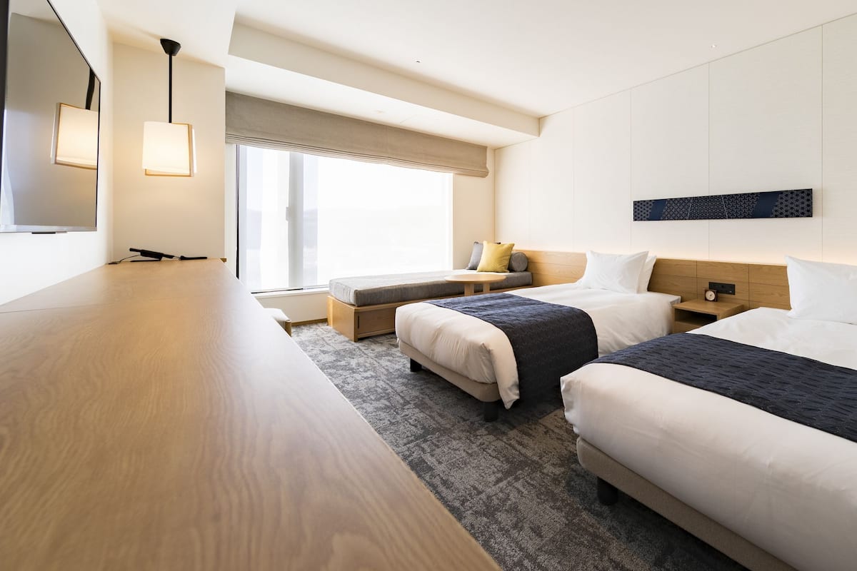 A modern hotel room with two beds, a wooden desk, a large window with a view, and a wall-mounted artwork.