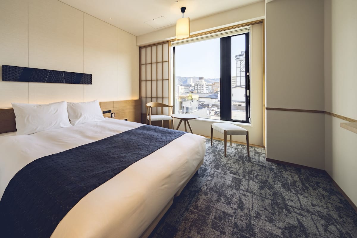 A hotel room with a large bed, a desk, a chair, and a view of the city through a large window.