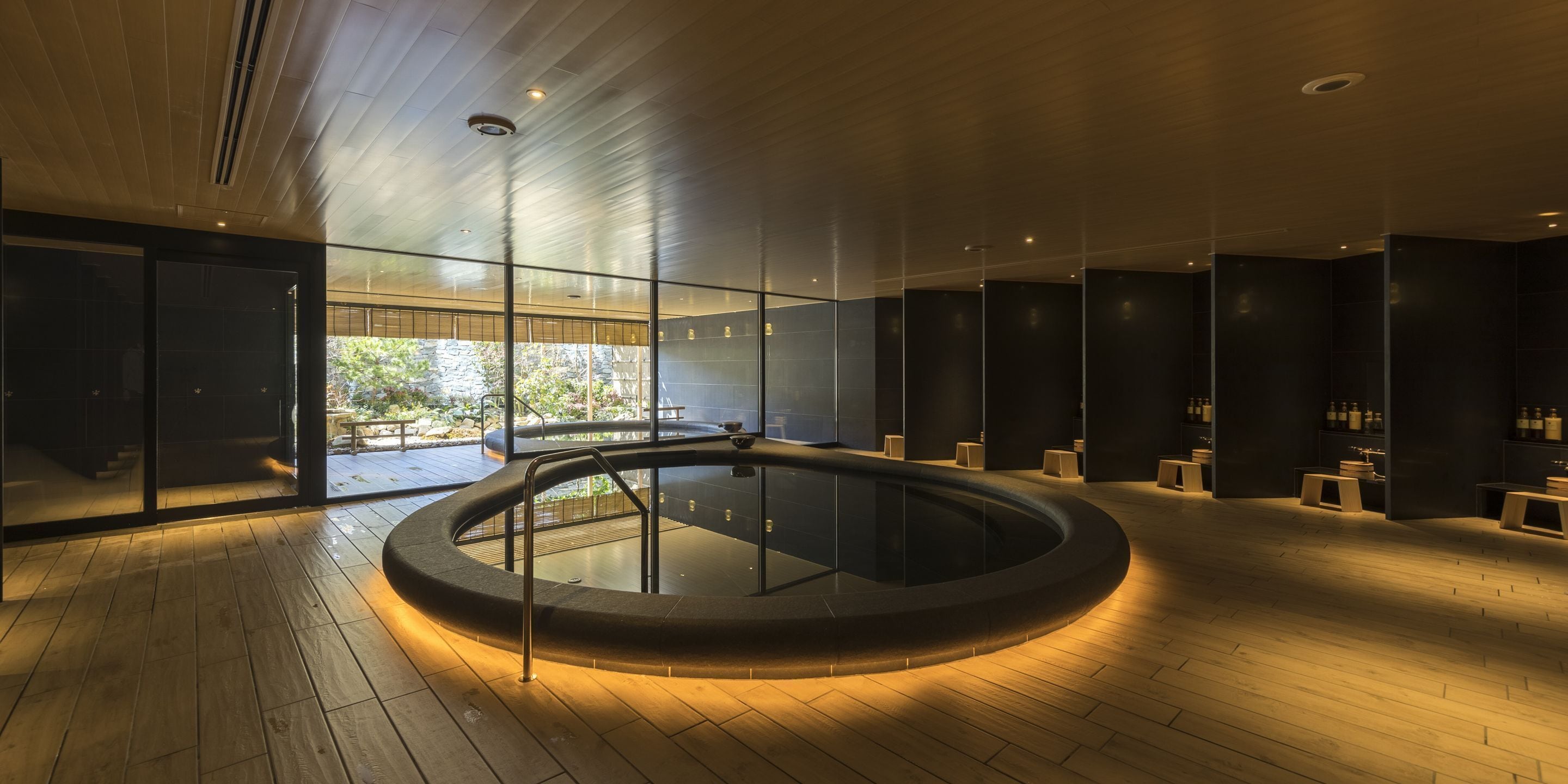 Uma área moderna de piscina interna com estrutura de vidro, piso de madeira e uma banheira de hidromassagem central.