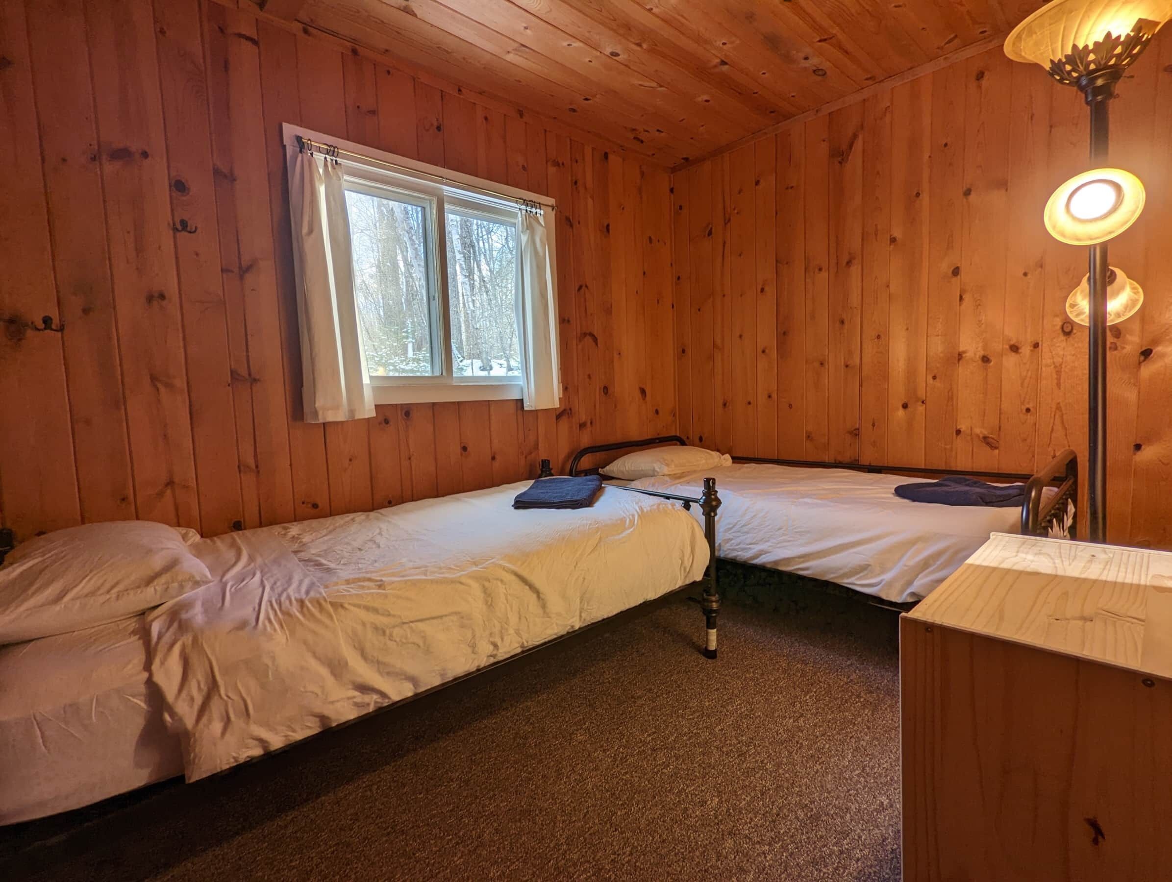 Une chambre de style cabane avec deux lits simples, une fenêtre avec des rideaux, un plafond en bois et une lampe de table.