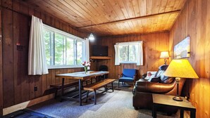 A cozy cabin interior with wooden walls, a dining table, a sofa, and two windows with curtains. - Premium Amenities Included! Cabin 9 - POV Resort (Phelps)