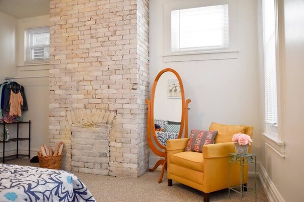 Une chambre avec un mur en briques, un fauteuil jaune, un miroir en bois et un lit avec une literie à motifs.