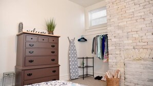 A bedroom with a bed, a dresser, a hanging rack with clothes, a brick wall, and a window. - Bright 1BR Retreat | Near Downtown & Airport (Pittsburgh)