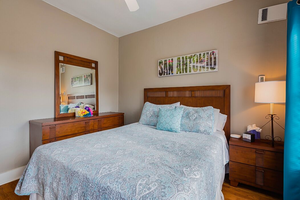 A bedroom with a bed, a dresser, a mirror, a wall-mounted picture, and a bedside table with a lamp.