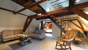 A loft-style living room with exposed wooden beams, a sofa, a rocking chair, and a staircase leading to an upper level.