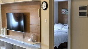 Una moderna camera d'albergo con un'ampia televisione a schermo piatto montata su un pannello di legno, un comodino con oggetti decorativi, una sveglia e un letto con biancheria a motivi.