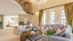 A spacious living room with a high ceiling, a chandelier, and a large window with curtains. There are two sofas, a coffee table, and a small side table. - Uber Luxurious NYC-Style Penthouse, Two Minutes Walk from Long Street (Cape Town)
