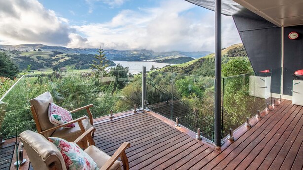 Une terrasse en bois dotée d’une balustrade en verre, offrant une vue panoramique sur un paysage pittoresque composé de collines et d’un lac.