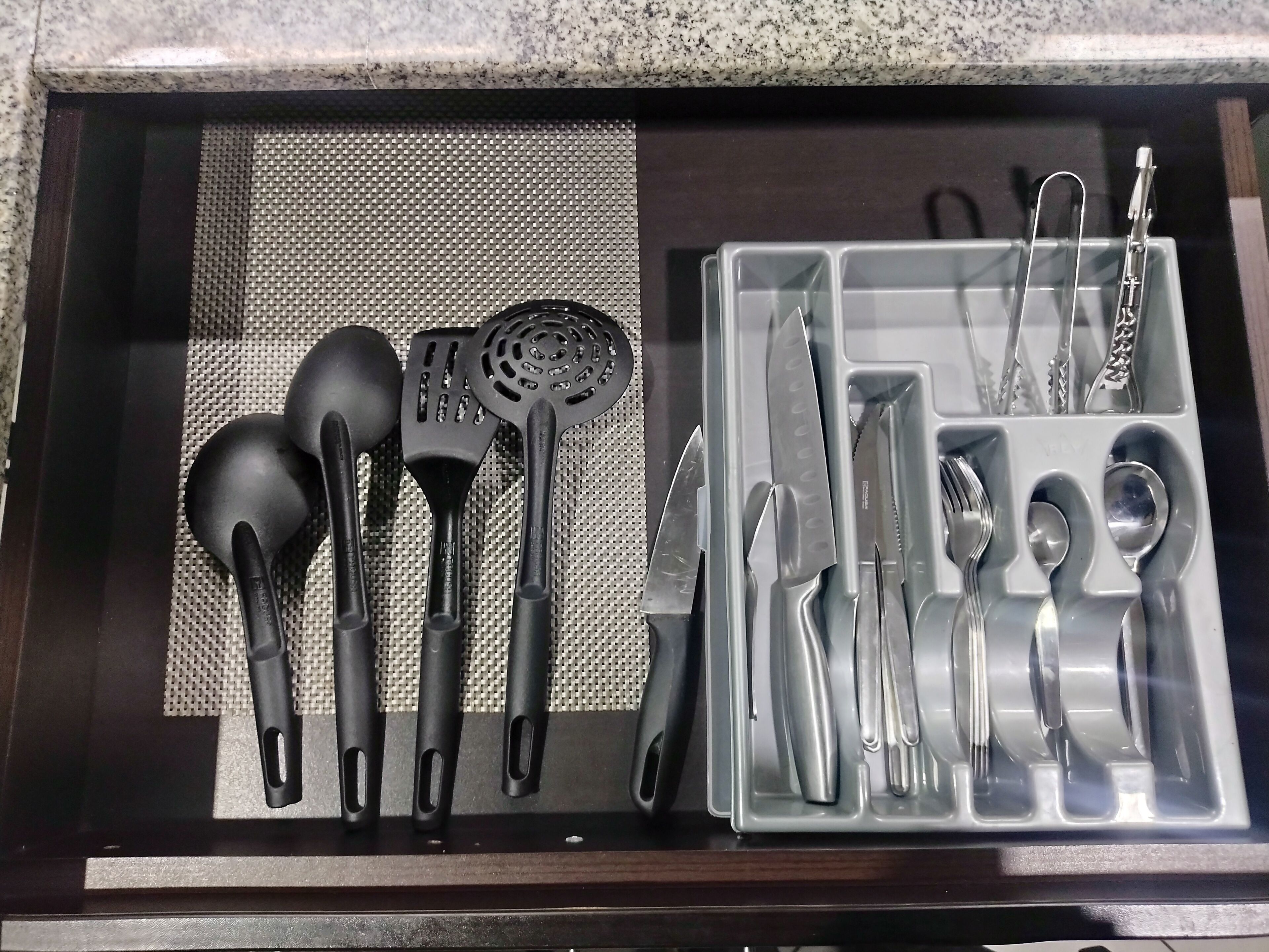 Un cajón de cocina con utensilios negros y grises, incluyendo espátulas, una cuchara escurridora y un juego de cuchillos.
