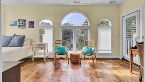A room with a bed, two wicker chairs with cushions, a small table with a lamp, and large arched windows with a view of the outside.