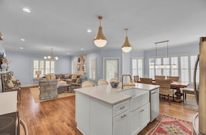 A spacious living room with a large island, a dining area with wicker chairs, and a kitchen with white cabinets and a central island.