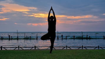 A person performing a yoga pose on a grassy area near a body of water with a sunset in the background.