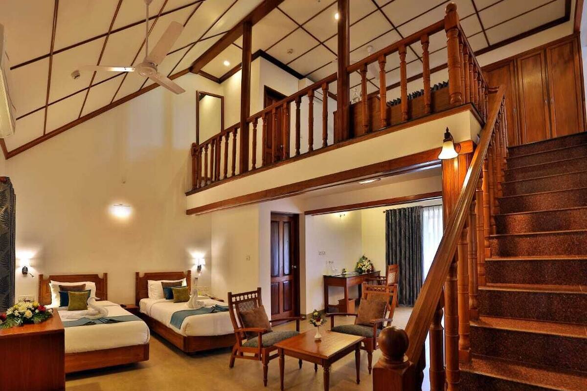 A spacious hotel room with a high ceiling, a wooden staircase, a balcony, and a dining area with a table and chairs.