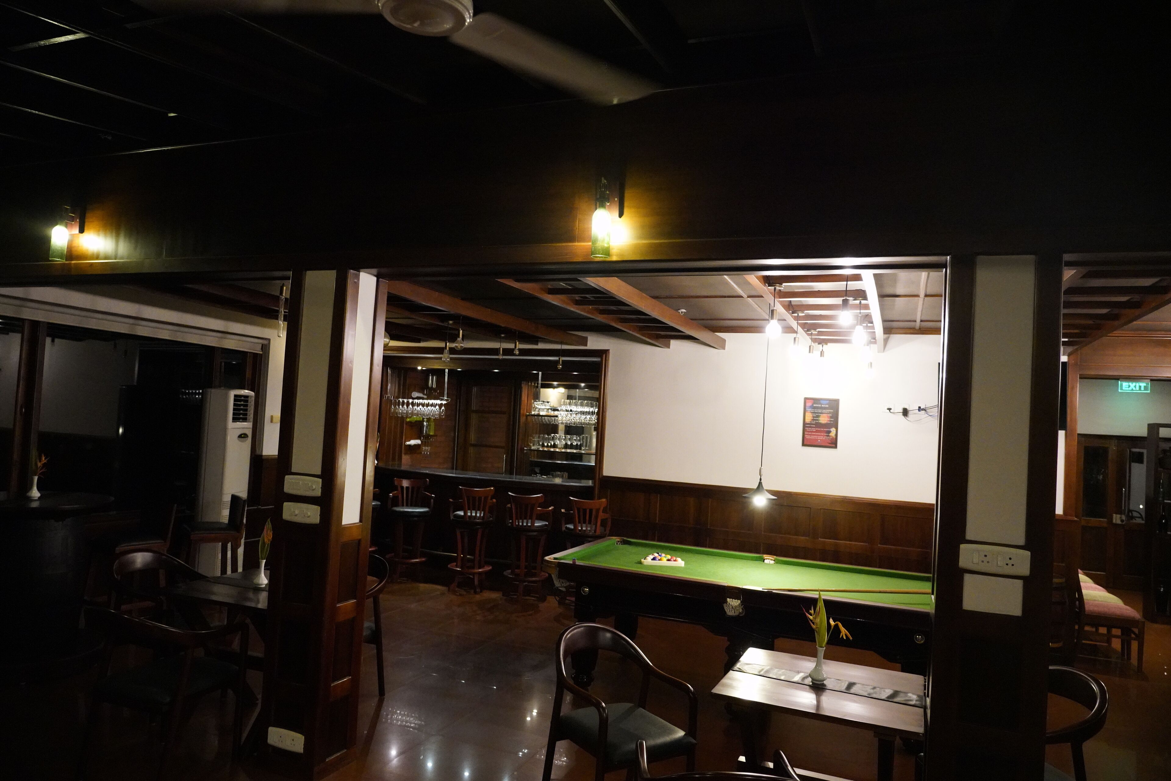 Zona de bar tenuemente iluminada con mesa de billar, sillas de madera y barra con vasos.