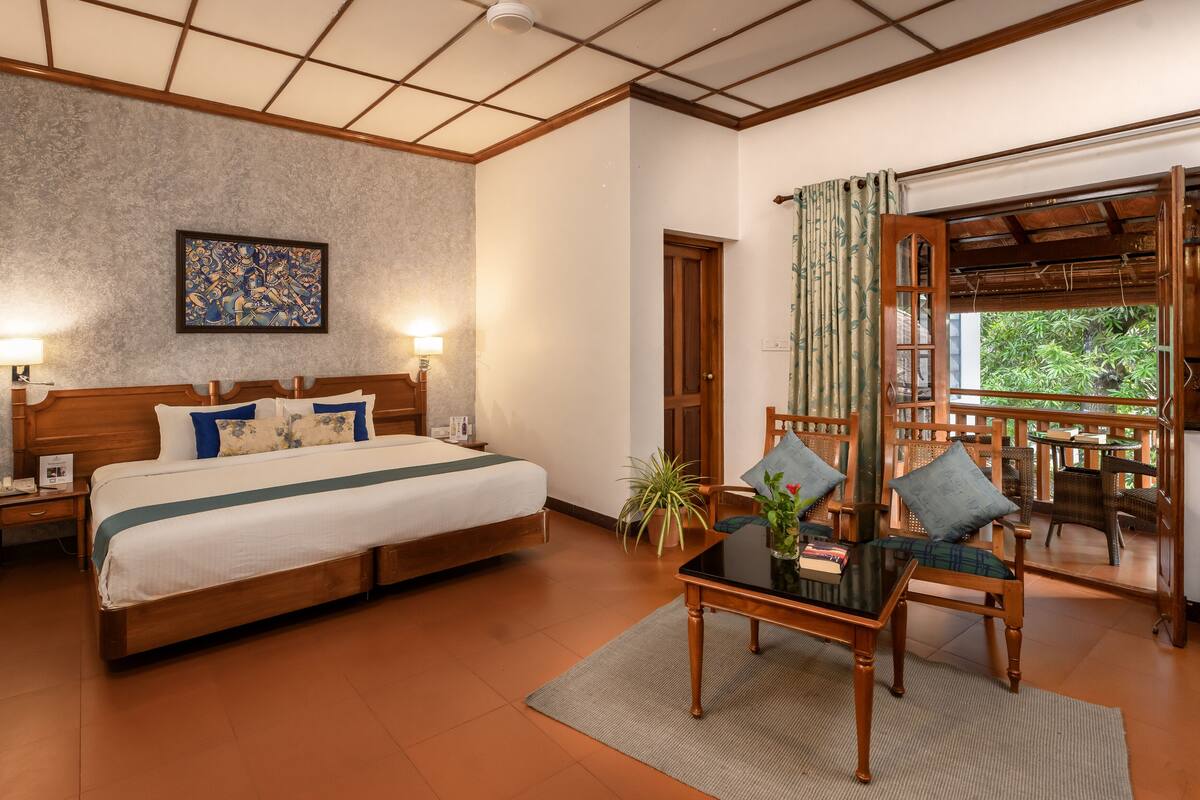 A hotel room with a wooden bed, two bedside lamps, a framed artwork, a balcony with wicker furniture, and a view of greenery.