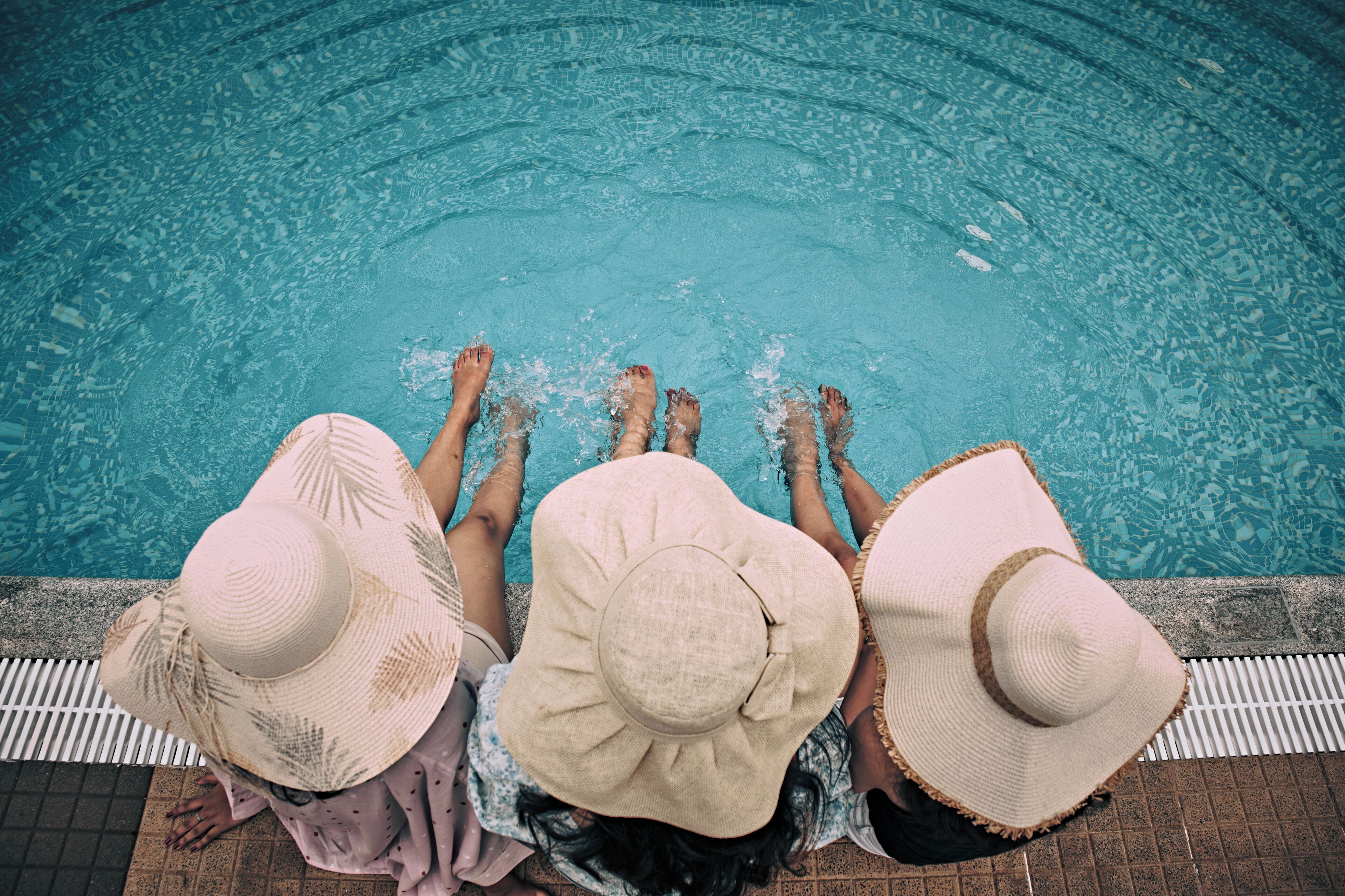 Three people sitting by a pool with their feet in the water, wearing hats.