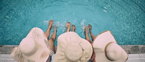 Three people sitting by a pool with their feet in the water, wearing hats.