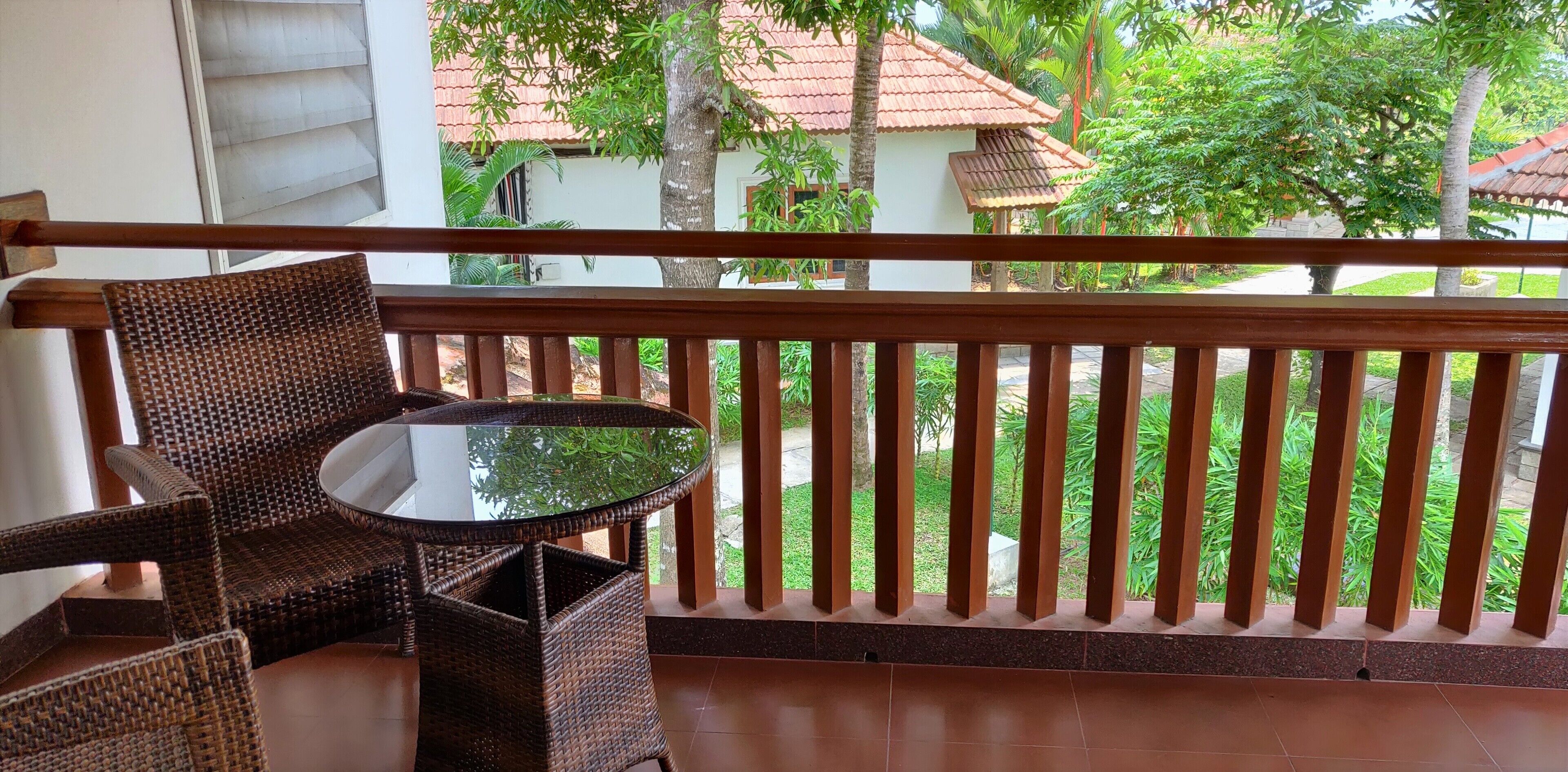 A balcony with wicker furniture and a glass table, overlooking a garden with trees and a building with a red roof.