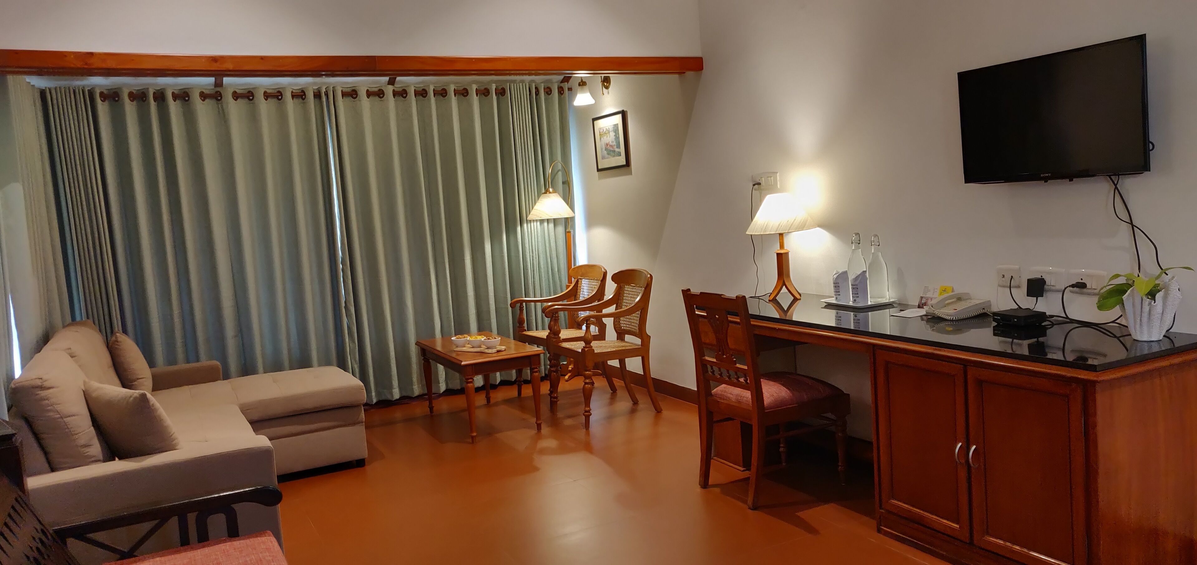 A hotel room with a sofa, two wooden chairs, a small table with a lamp, a TV, and a wooden cabinet.
