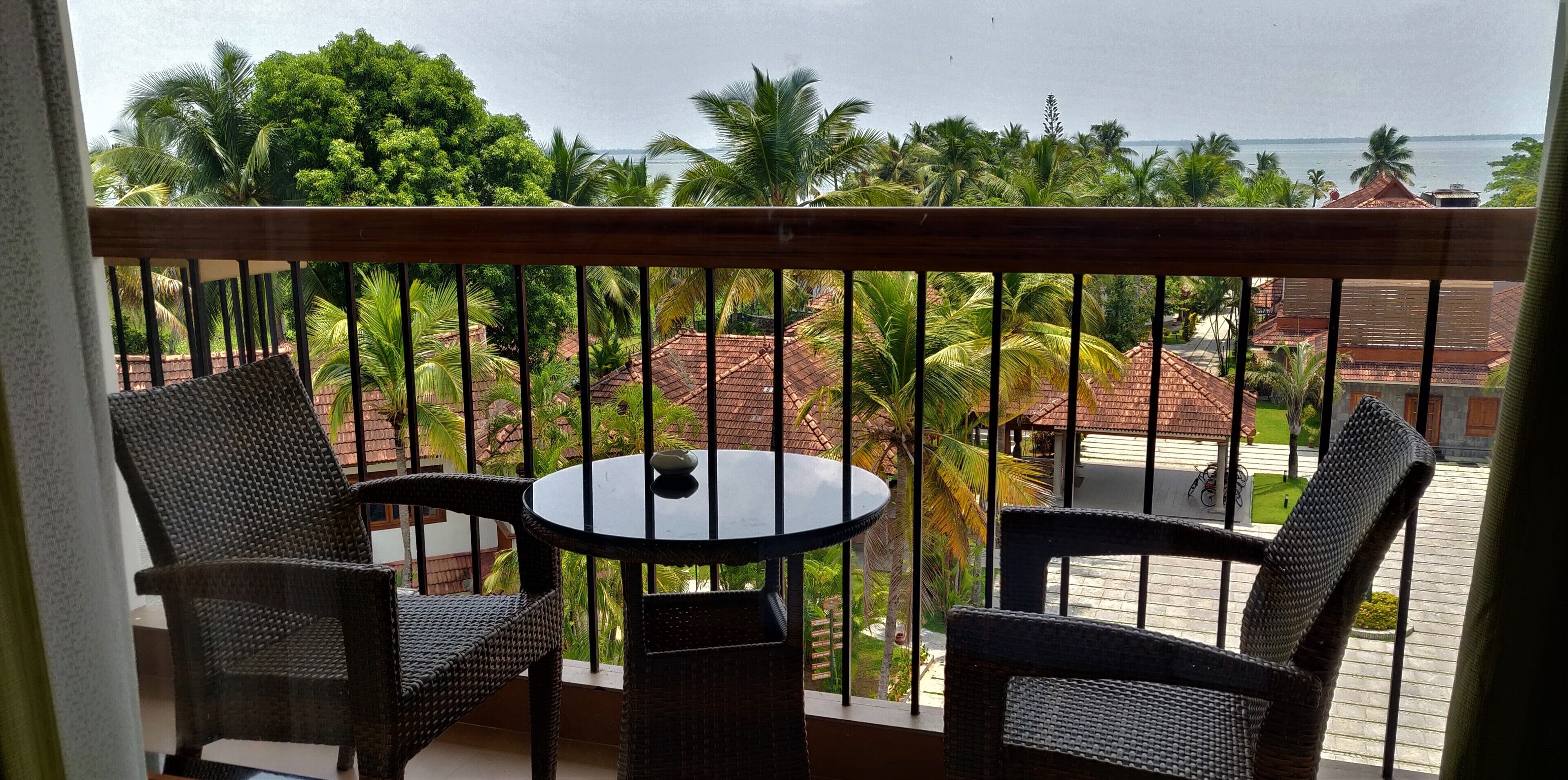 A balcony with wicker furniture, a round table, and a view of a tropical landscape with palm trees and a building with a red roof.