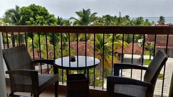A balcony with wicker furniture, a round table, and a view of a tropical landscape with palm trees and a building with a red roof.