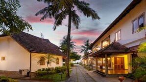 A pathway flanked by buildings with tiled roofs, palm trees, and a sign indicating a room number. - Backwater Ripples Kumarakom (Kottayam)