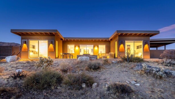 A modern two-story building with large windows and doors, a patio area with outdoor seating, and a desert landscape. - Shelter 2 By Homestead Modern (Joshua Tree)