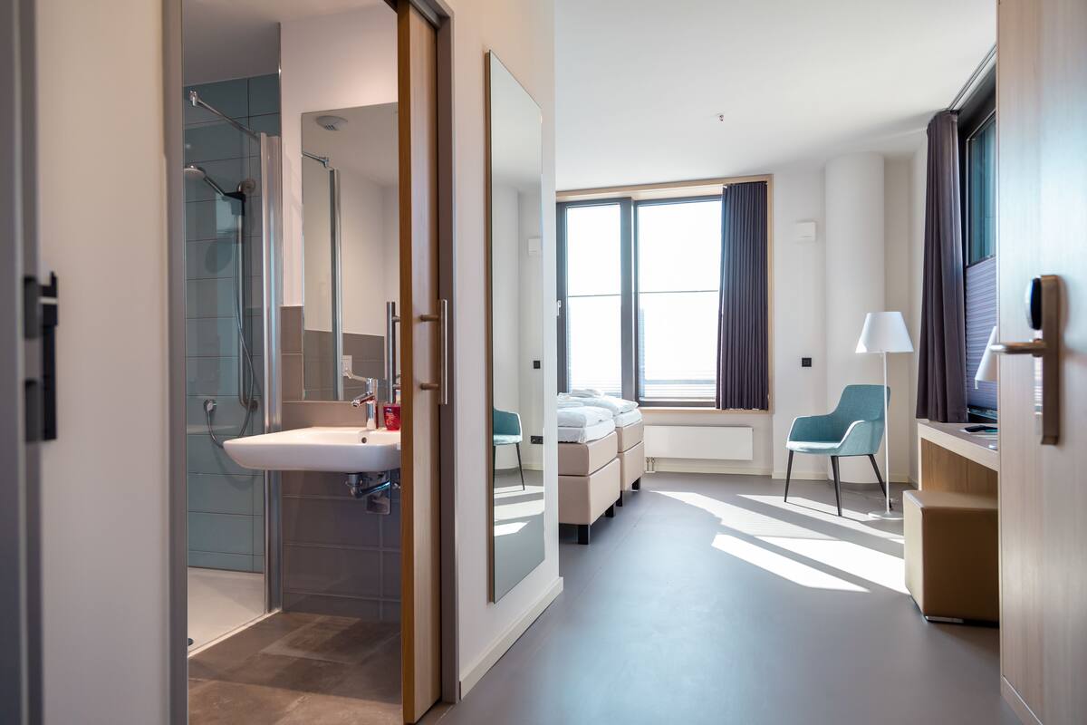 A modern hotel room with a glass shower, a large mirror, a bed with a nightstand, a chair, and a TV.