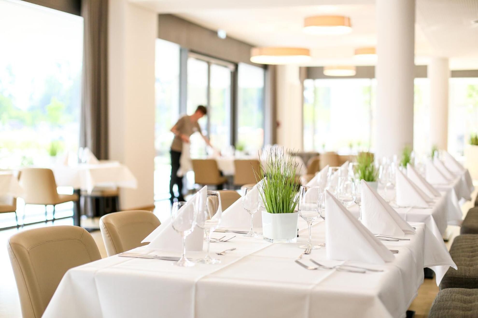 A restaurant with long tables set for dining, a waiter in the background, and large windows allowing natural light.