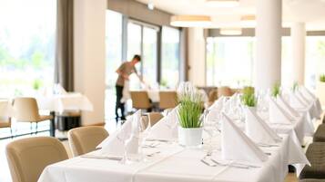 A restaurant with long tables set for dining, a waiter in the background, and large windows allowing natural light.
