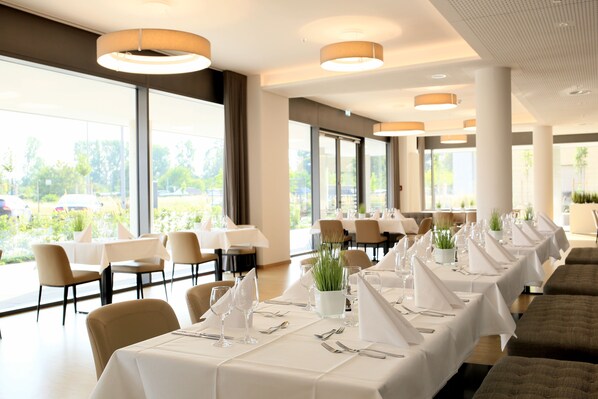 A restaurant with long tables set for dining, large windows, and indoor plants.