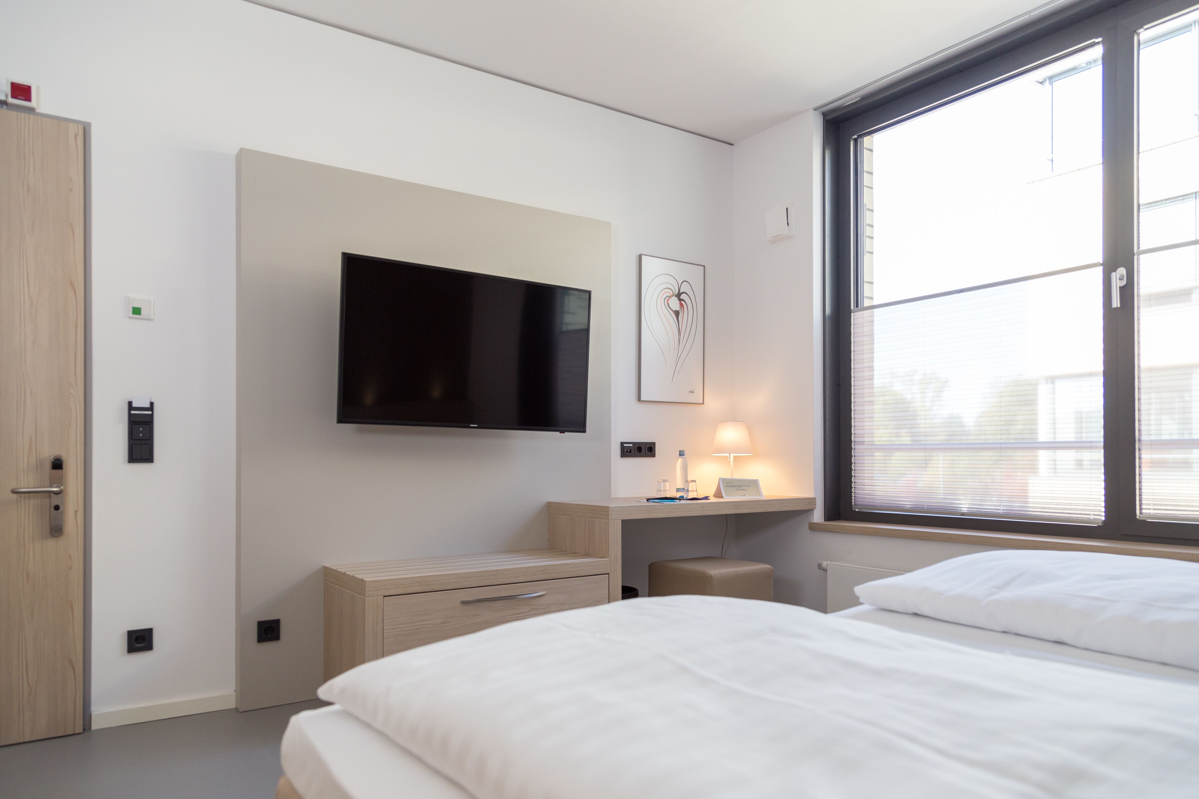Ein modernes Hotelzimmer mit einem großen Flachbildfernseher an der Wand, einem Nachttisch mit Lampe und einem Fenster mit Jalousien.