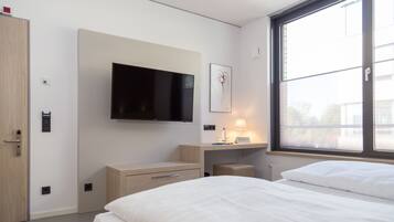 Ein modernes Hotelzimmer mit einem großen Flachbildfernseher an der Wand, einem Nachttisch mit Lampe und einem Fenster mit Jalousien.