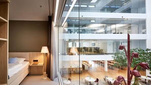 Un hotel moderno con cama, mesita de noche, lámpara y vistas al comedor interior del hotel a través de un muro de cristal.