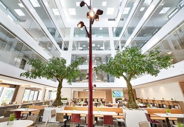 A modern indoor dining area with high ceilings, large windows, and two central trees. - Hotel Vivendi (Paderborn)