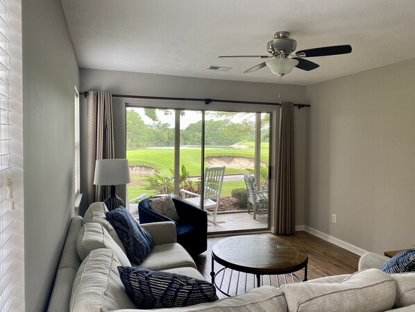 A modern living room with a sofa, chairs, a table, and a view of a golf course through large sliding glass doors. - Sunset on 17 at TrueBlue (Pawleys Island)