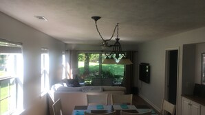Ein Essbereich mit einem Tisch für vier Personen, Stühlen, einer Deckenleuchte und Blick auf einen Garten durch eine Schiebetür.