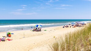 Una playa con gente relajándose bajo sombrillas, algunos sentados en sillas y otros caminando por la orilla.