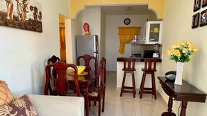 A living room with a sofa, dining table, chairs, a kitchen area with a refrigerator, and a small table with a vase of flowers. - Higuey Center City, Candyland 2 bedroom  (Higüey)