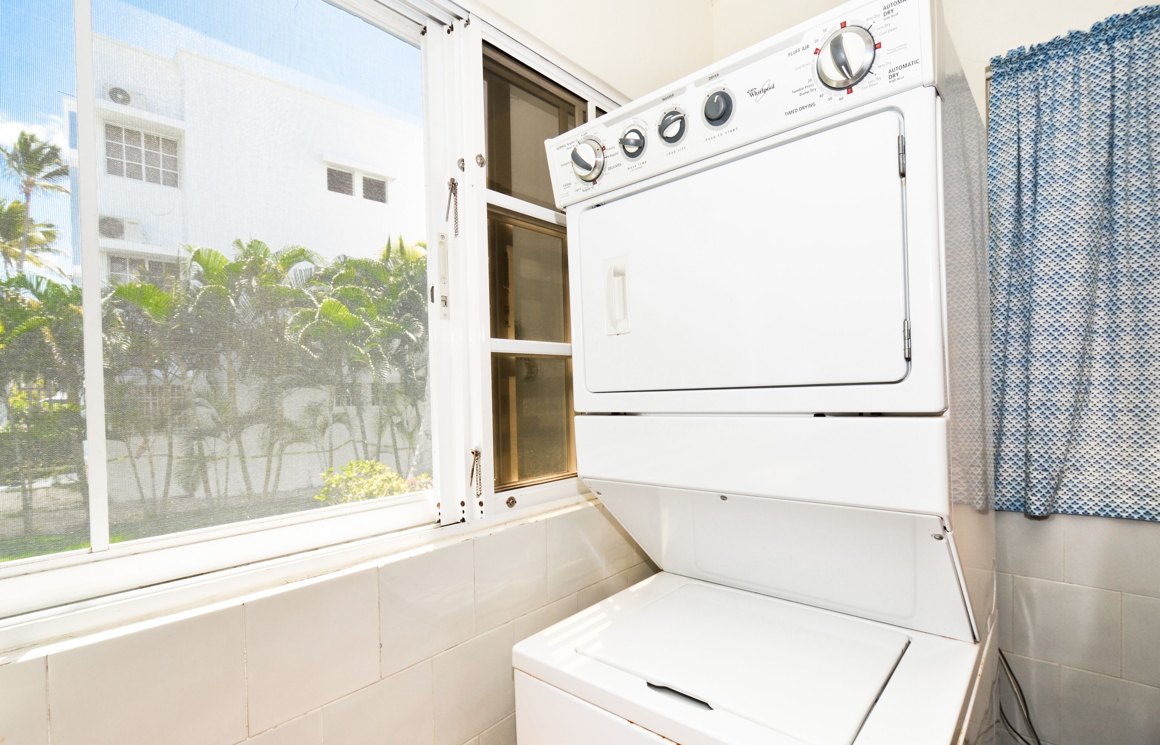 A laundry room with a stacked washer and dryer, a window with a view of palm trees, and a blue patterned curtain.