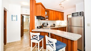 Eine Küche mit Holzausstattung, einem Edelstahlkühlschrank, einer Mikrowelle und einer Arbeitsplatte aus Granit. Zwei weiße Barhocker mit blauen Kissen sind vorhanden.